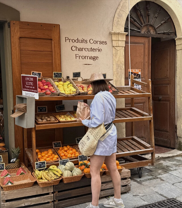 Manon devant une épicerie corse, panier en osier à l'épaule